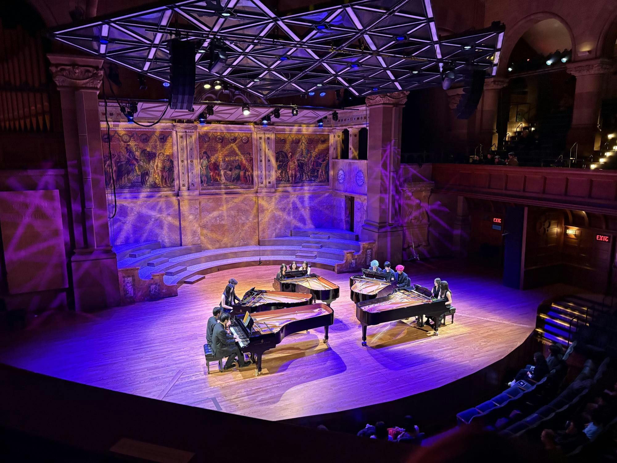 Five pianos with twelve pianists seated around them. The stage is lit in blue-purple light.