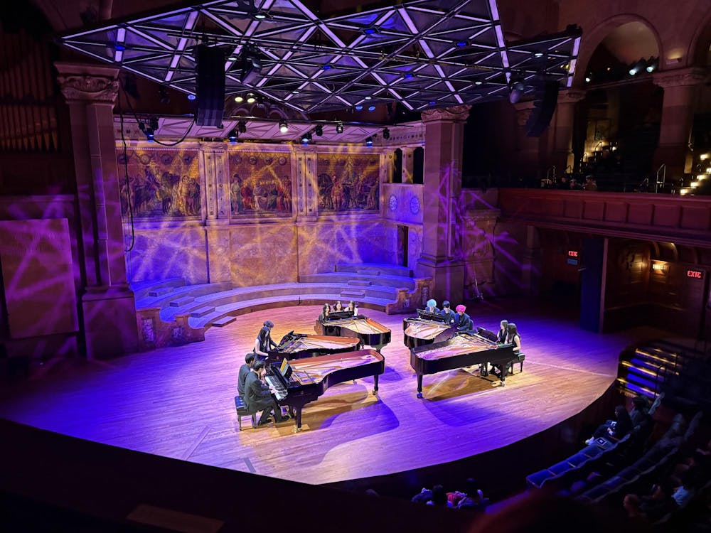 Five pianos with twelve pianists seated around them. The stage is lit in blue-purple light.