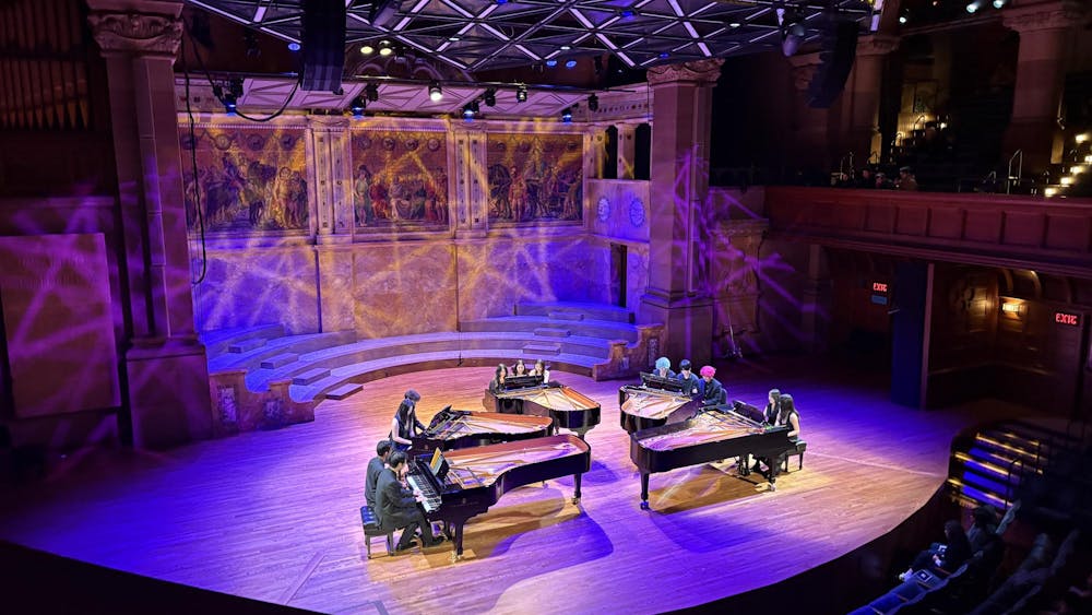 Five pianos with twelve pianists seated around them. The stage is lit in blue-purple light.