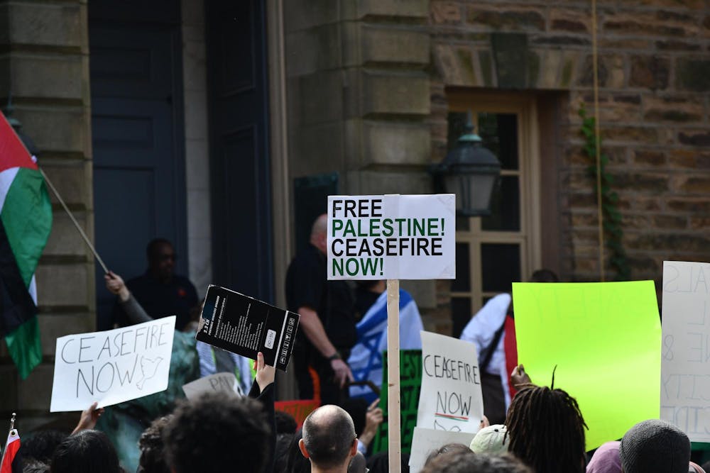 A number of people hold up signs reading "Free Palestine Ceasefire Now" and "Ceasefire Now" holding Palestinian flags. A person holding an Israeli flag is visible in the background.