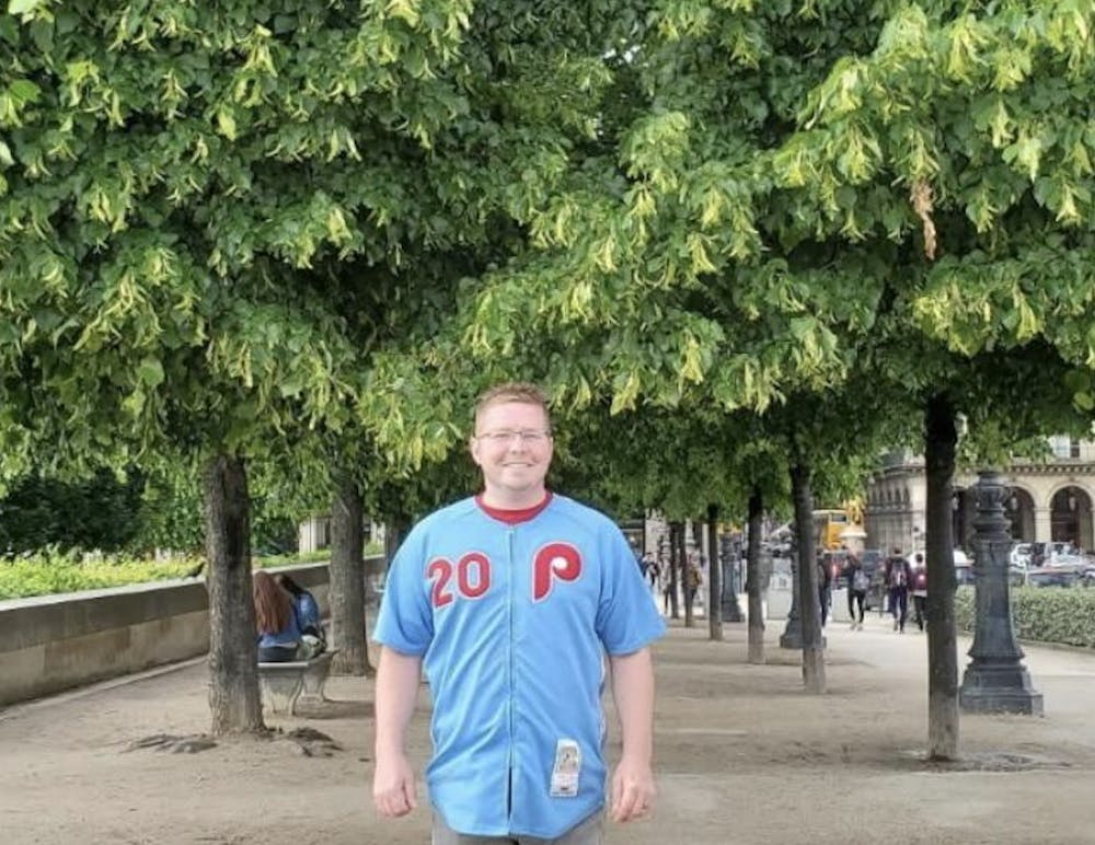 A man in a light blue jersey with red text and wearing glasses stands before a double row of trees with large, waxy green leaves. 