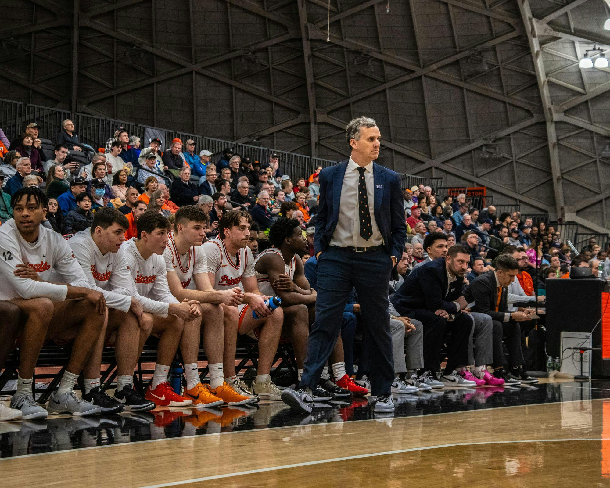 A man wearing a blue suit with a gray button down walking across the baseline as a group of coaches and players sit behind him with fans watching in the background.
