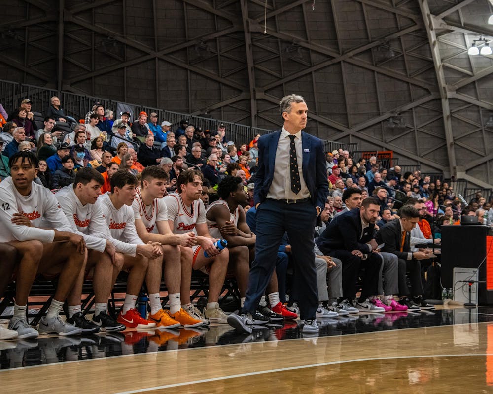 A man wearing a blue suit with a gray button down walking across the baseline as a group of coaches and players sit behind him with fans watching in the background.