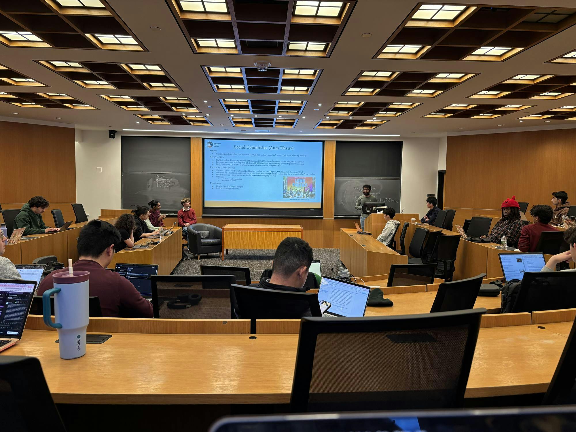 A room with wooden desks. A few people are set at them with laptops. Someone is stood at the front of the room presenting.
