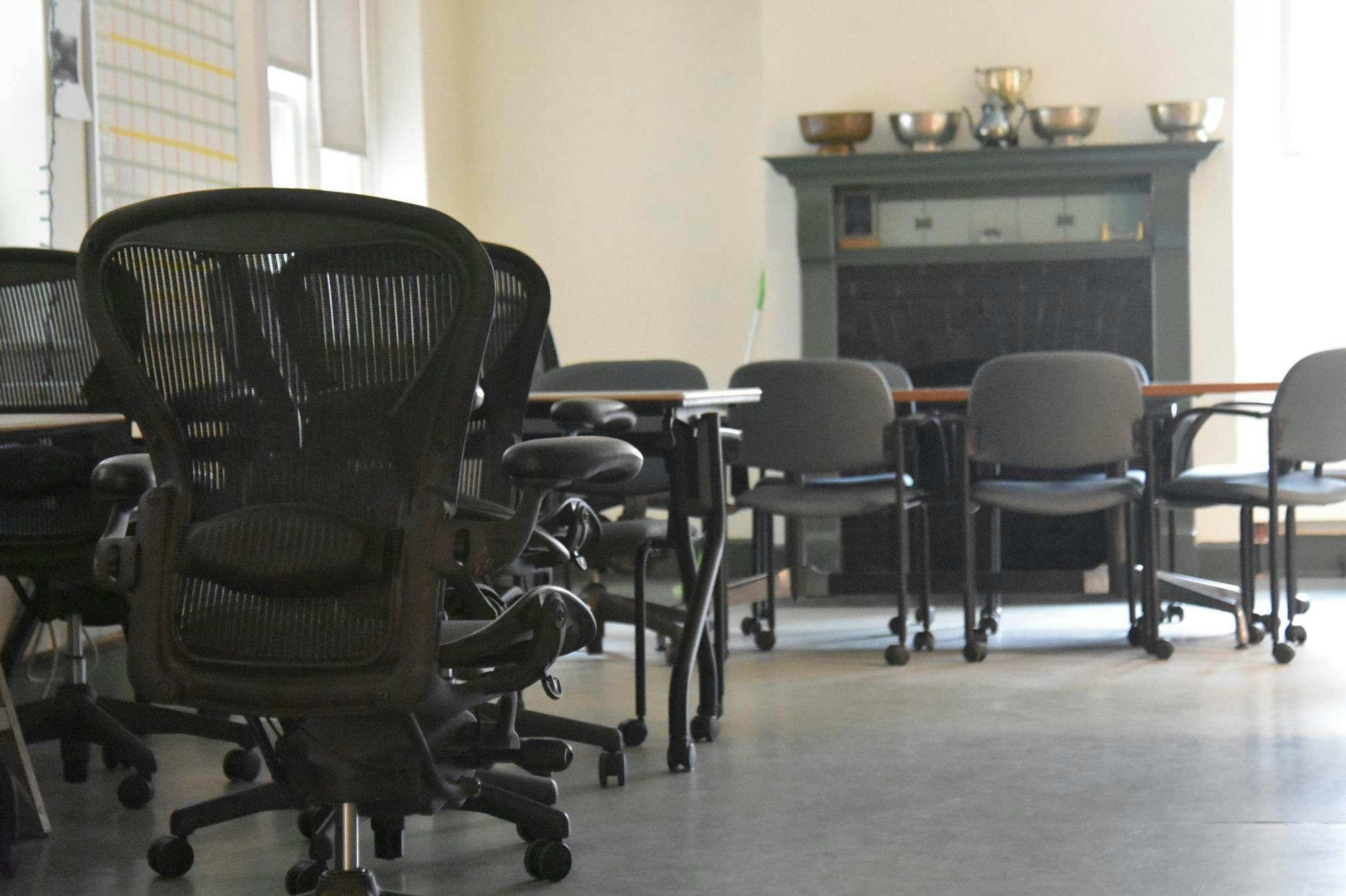 A black desk chair with light shining through the mesh backing in the foreground; in the background, a brown table surrounded by chairs in front of a green fireplace.