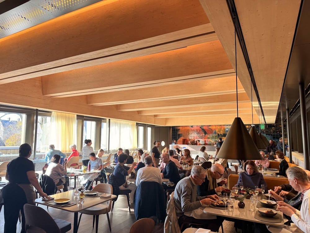 A large number of people sat inside a restaurant. Lights hang down from the ceiling while sunlight shines through the windows.