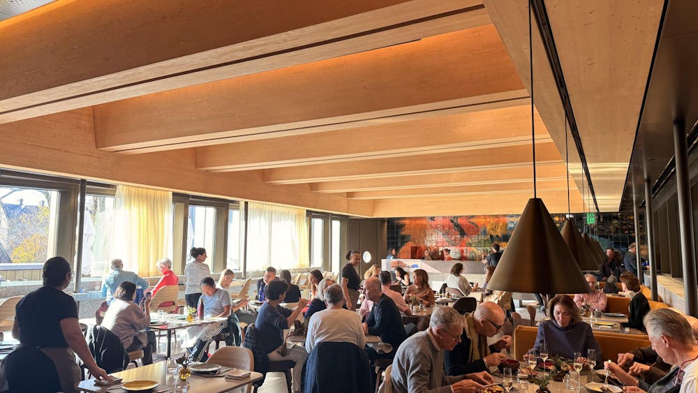 A large number of people sat inside a restaurant. Lights hang down from the ceiling while sunlight shines through the windows.