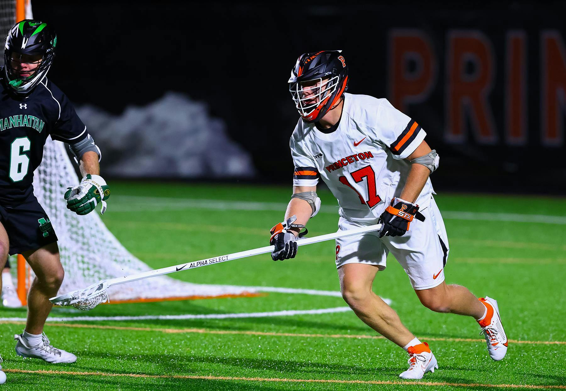 A man holding a lacrosse stick on a grass field with a goal in the background and a defender nearby.