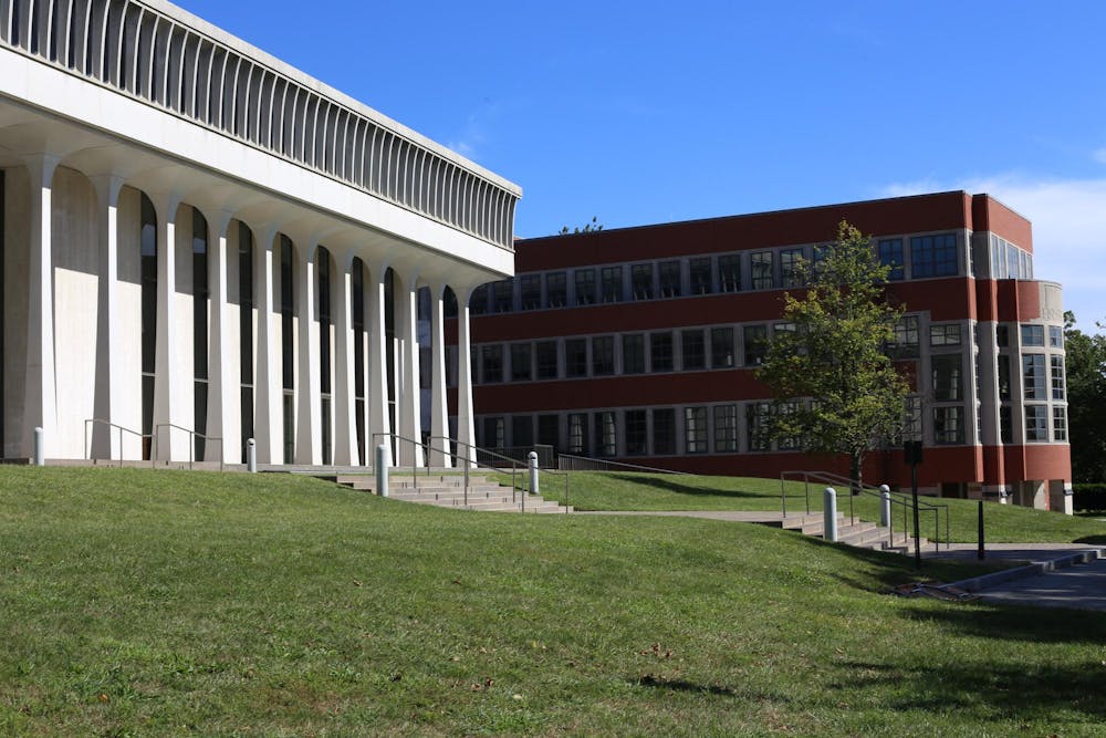 A white building with columns stands on a grassy hill in front of a brick building. 