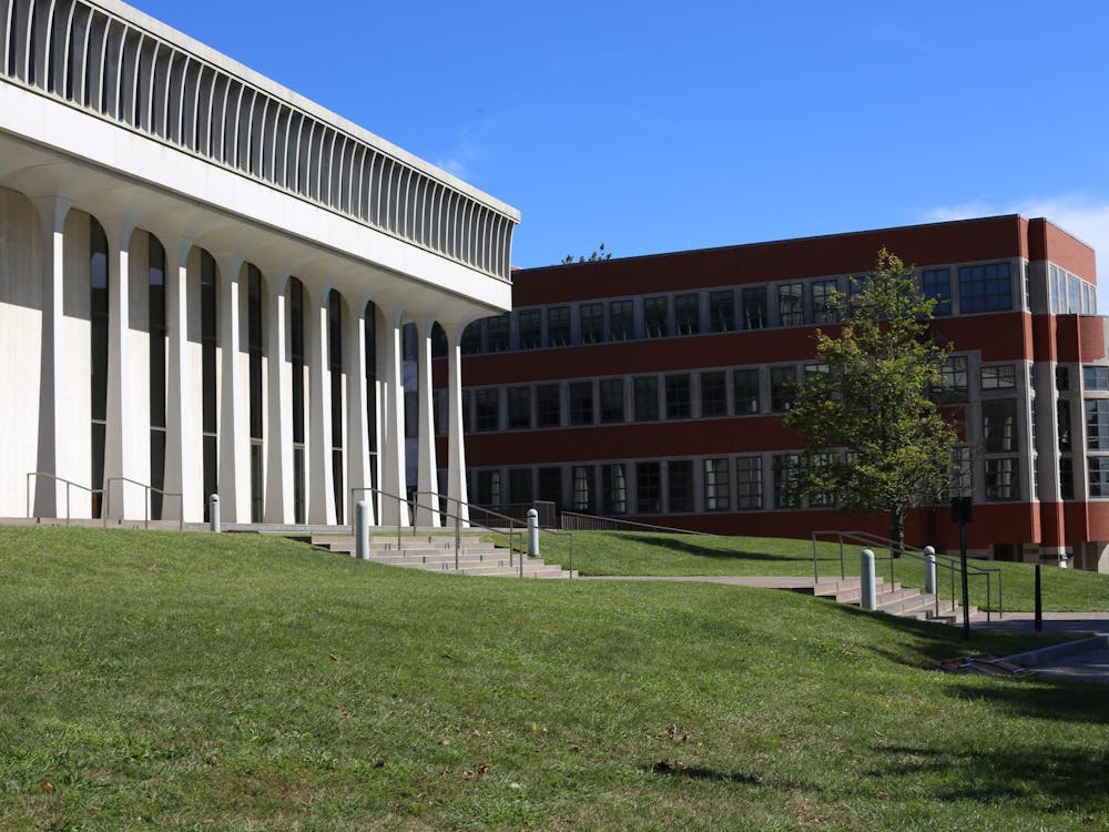 A white building with columns stands on a grassy hill in front of a brick building. 