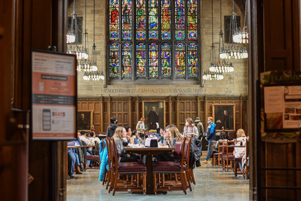 Several students pictured eating in the Graduate School dining hall in the center of the image, with the doors to the dining hall visible, but blurred in the foreground of the photo. 