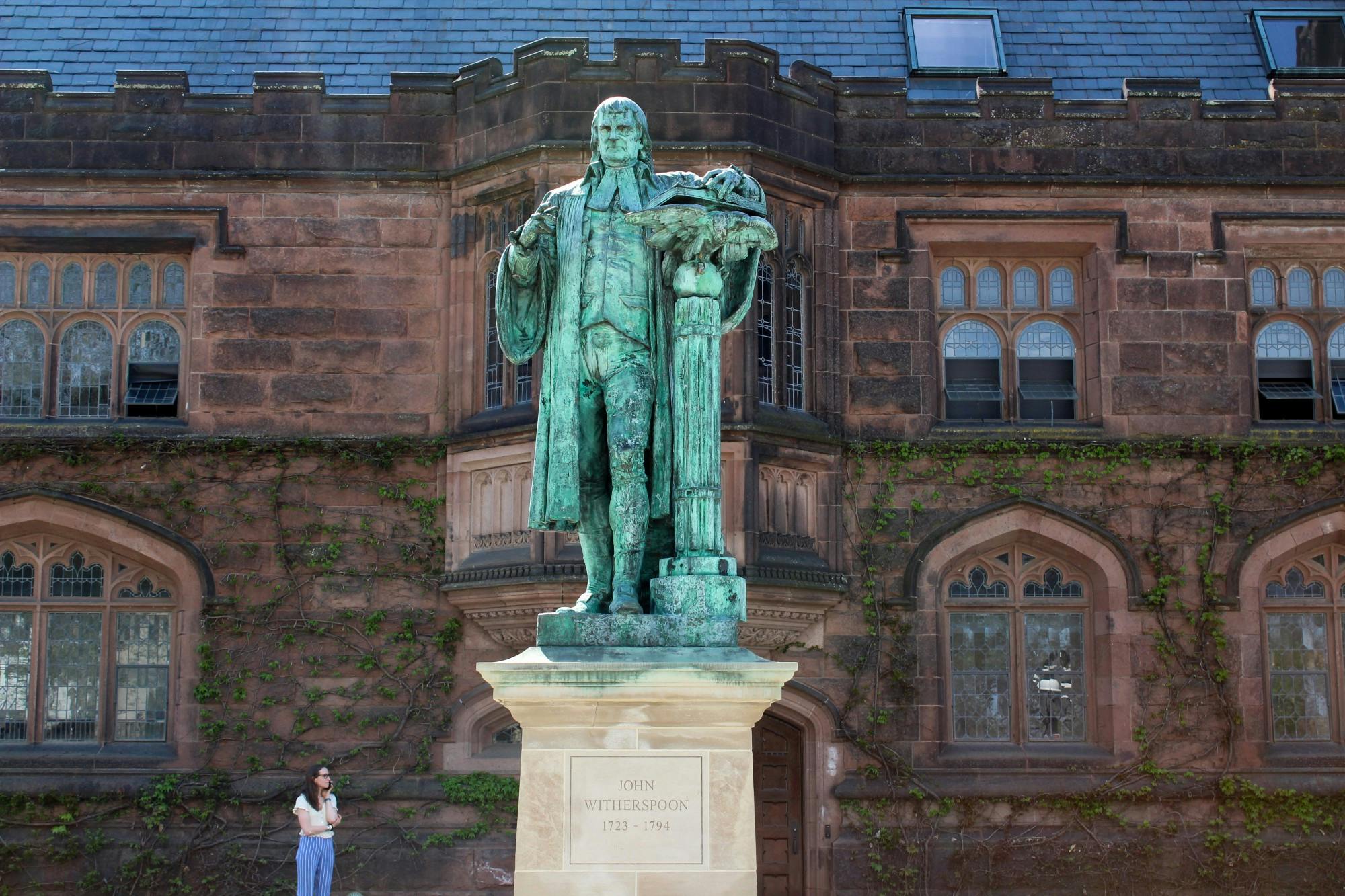 Oxidized bronze statue of a man on a stone pedestal in front of a neo-Gothic building. 
