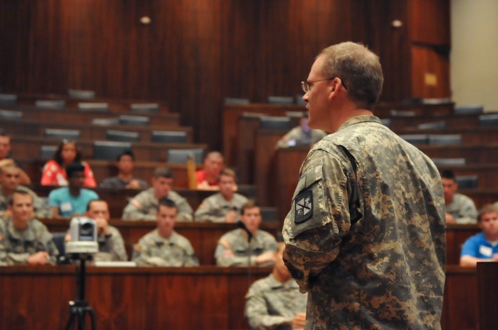 Man in camouflage uniform at a podium, speaking to an audience sitting in a wood-paneled room. 