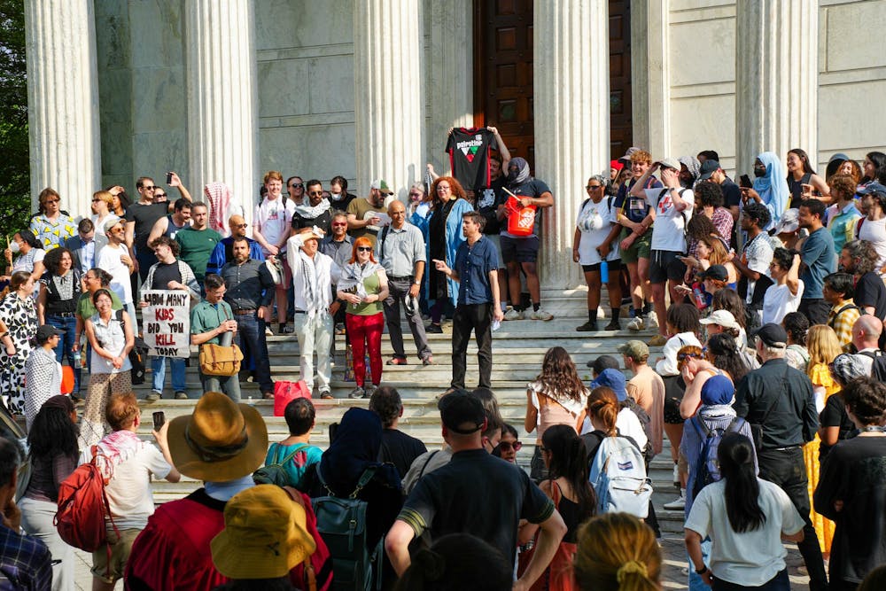A man holding a megaphone stands in the center of a crowd on the steps of a building with columns. 