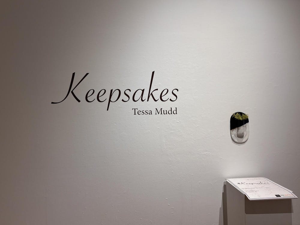 A white wall has black curvy text on it that reads "Keepsakes" in large text with a smaller "Tessa Mudd" to the bottom right of the title.