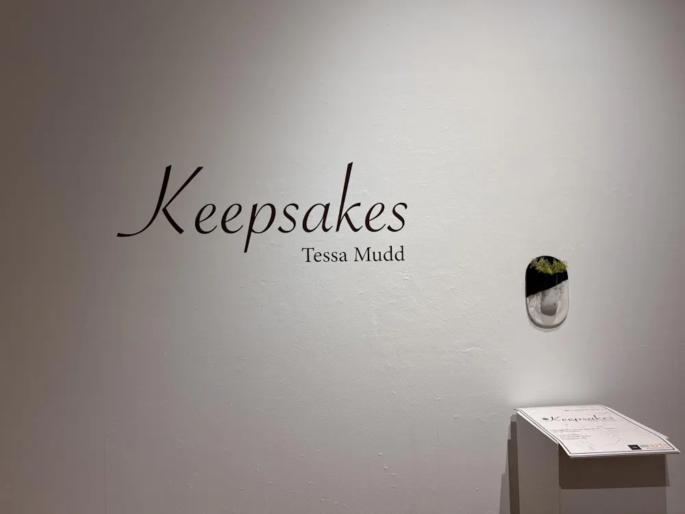 A white wall has black curvy text on it that reads "Keepsakes" in large text with a smaller "Tessa Mudd" to the bottom right of the title.