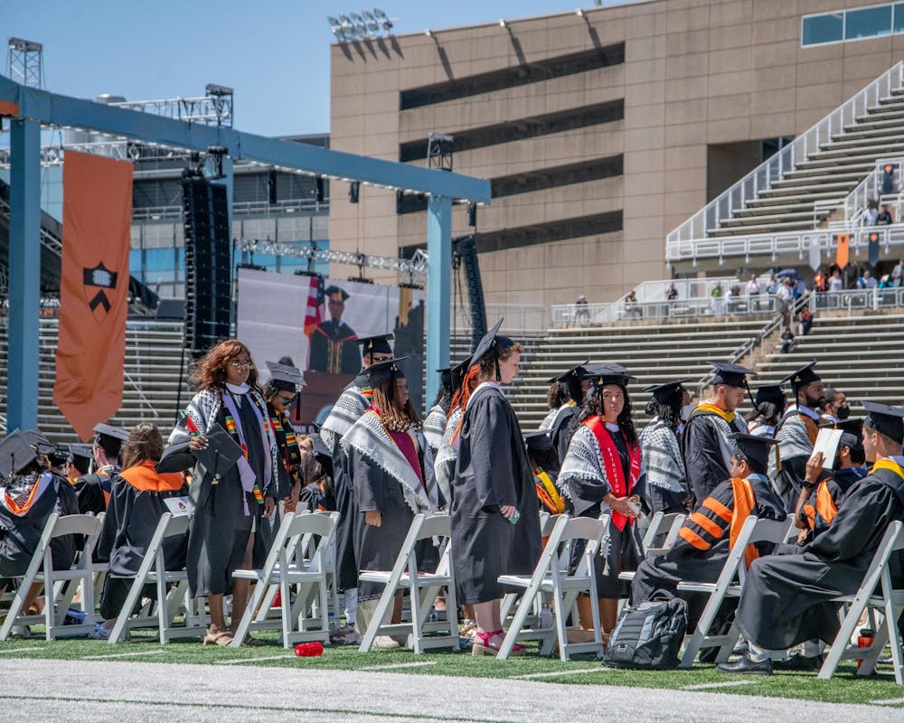 Around 15 students, many wearing keffiyehs, are facing away from the stage. Other graduates are seated and looking forward. In the background, President Eisgruber is shown on a large screen giving a speech.