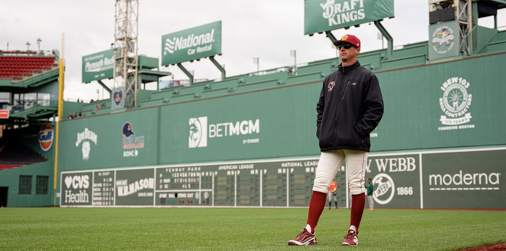 A man in a red and yellow hat, black quarter zip, and baseball pants stands on a baseball diamond.