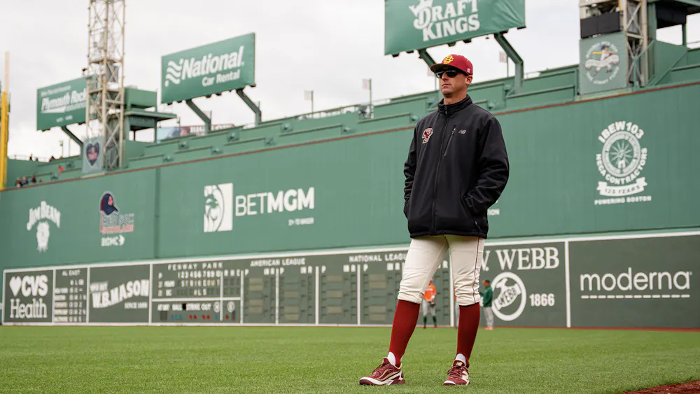 A man in a red and yellow hat, black quarter zip, and baseball pants stands on a baseball diamond.