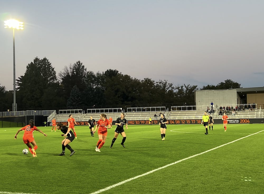 Soccer players in orange and black jerseys are scattered across the green in pursuit of the ball. 