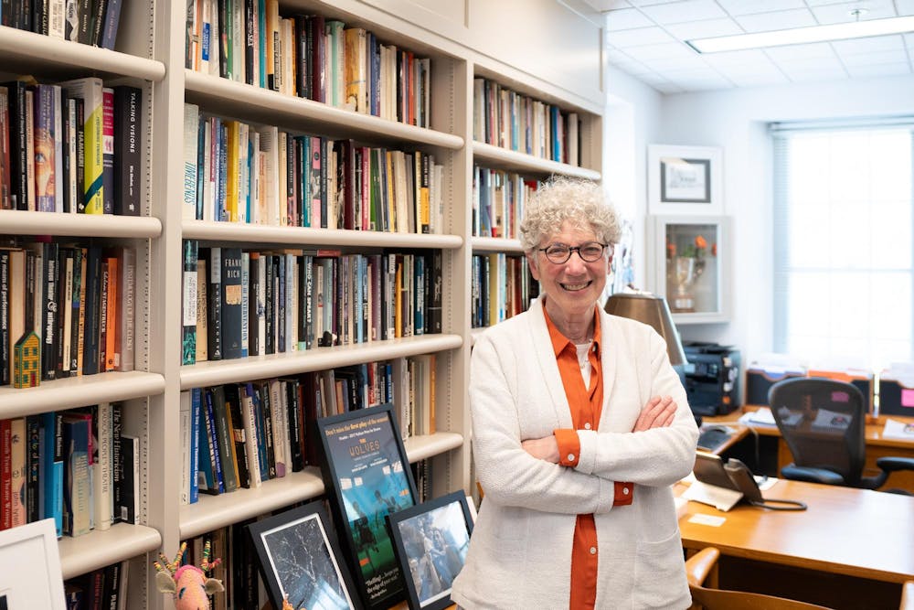 A white-haired woman with round glasses smiles in front of a large bookshelf in a brightly lit office.