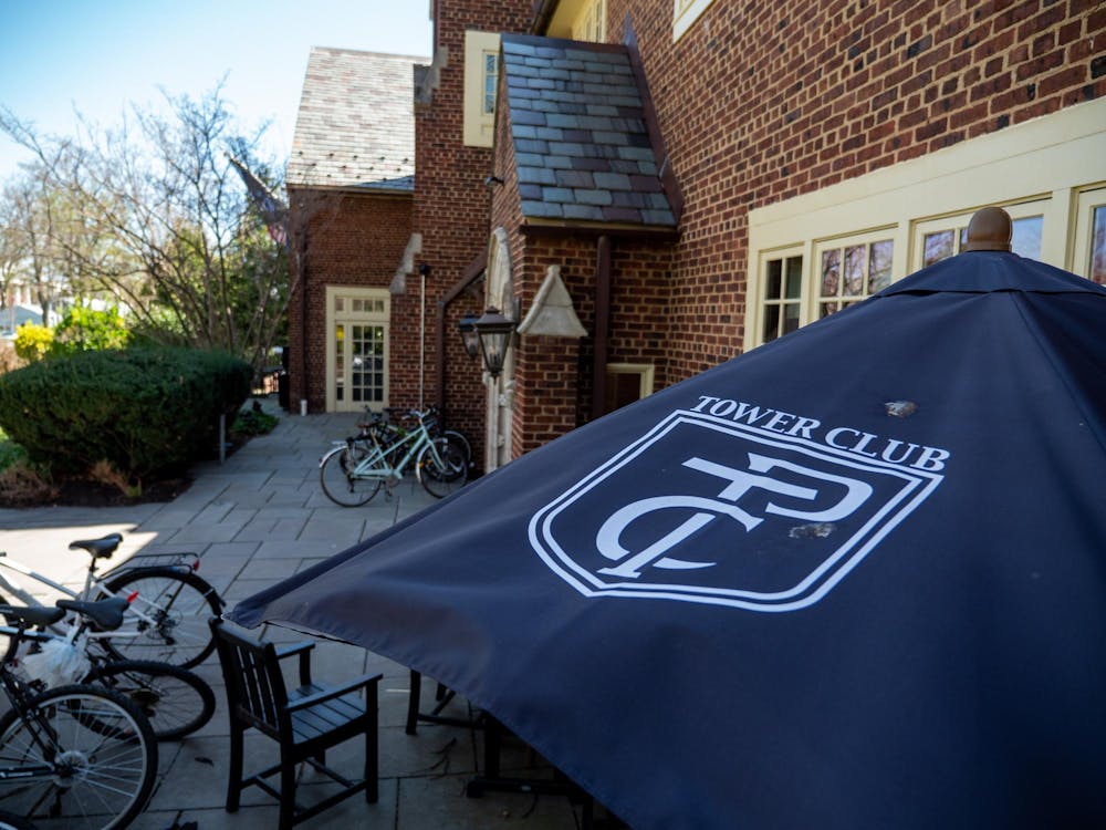 In the right foreground, a blue umbrella with text "TOWER CLUB" in white overtop and "PTC" insignia. In the background, a brown brick building with green bushes to the left.