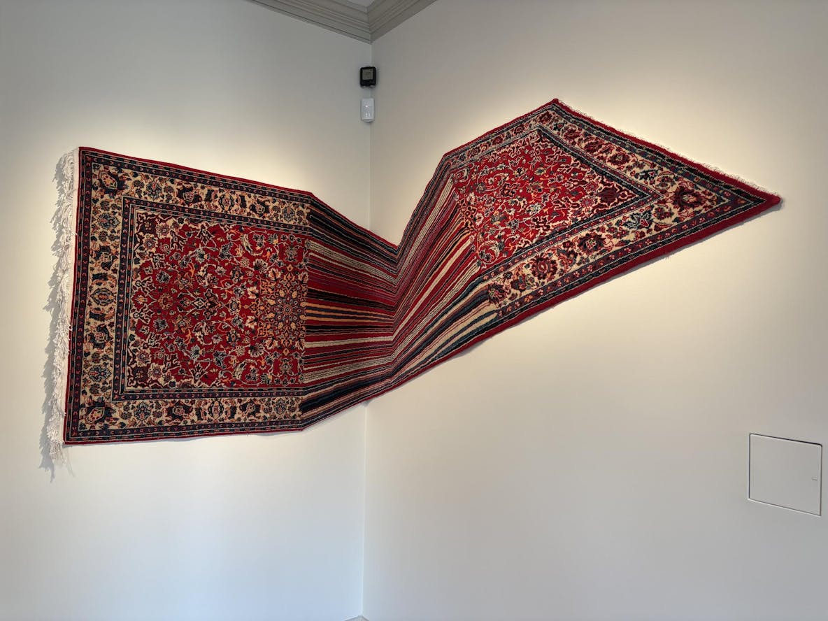 A red patterned carpet hangs between two walls, appearing almost as if being stretched and warped as it reaches the corner. 