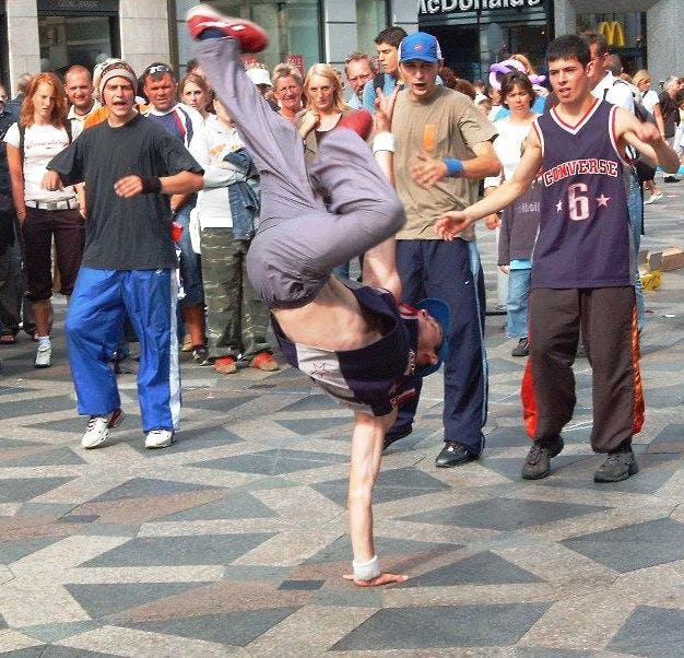 Breakdancer does a one-handed air freeze in front of three other b-boys with a small crowd behind in a public area in Copenhagen with a McDonald's in the background