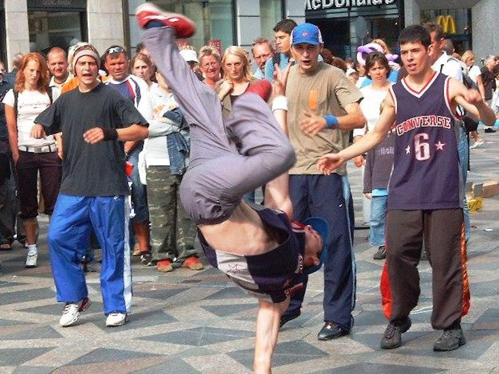 Breakdancer does a one-handed air freeze in front of three other b-boys with a small crowd behind in a public area in Copenhagen with a McDonald's in the background