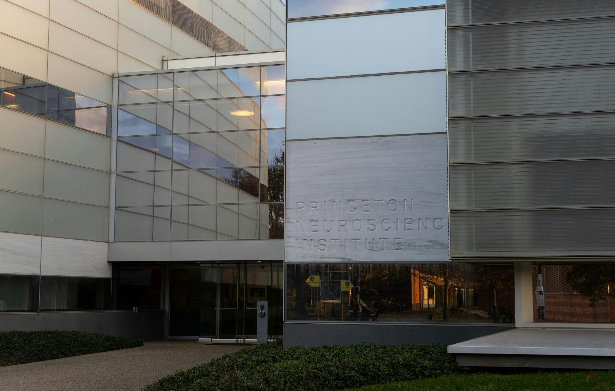 A large glass, metal and stone building that features a marble like stone sign that reads "Princeton Neuroscience Institute". The lighting is dim and the sun is setting.