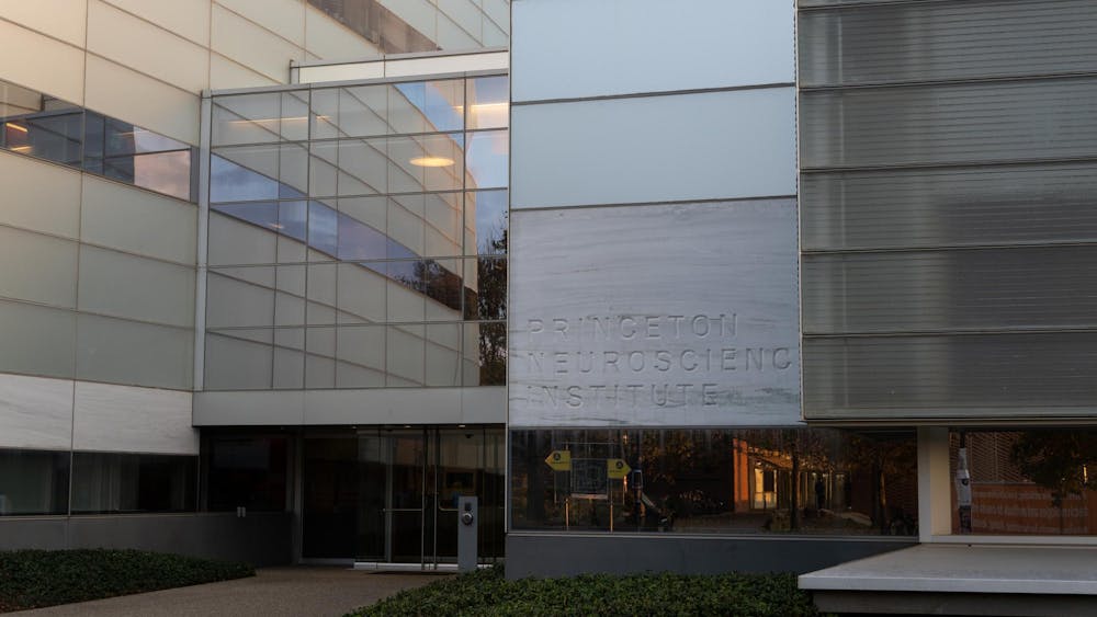 A large glass, metal and stone building that features a marble like stone sign that reads "Princeton Neuroscience Institute". The lighting is dim and the sun is setting.