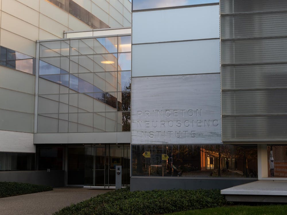 A large glass, metal and stone building that features a marble like stone sign that reads "Princeton Neuroscience Institute". The lighting is dim and the sun is setting.