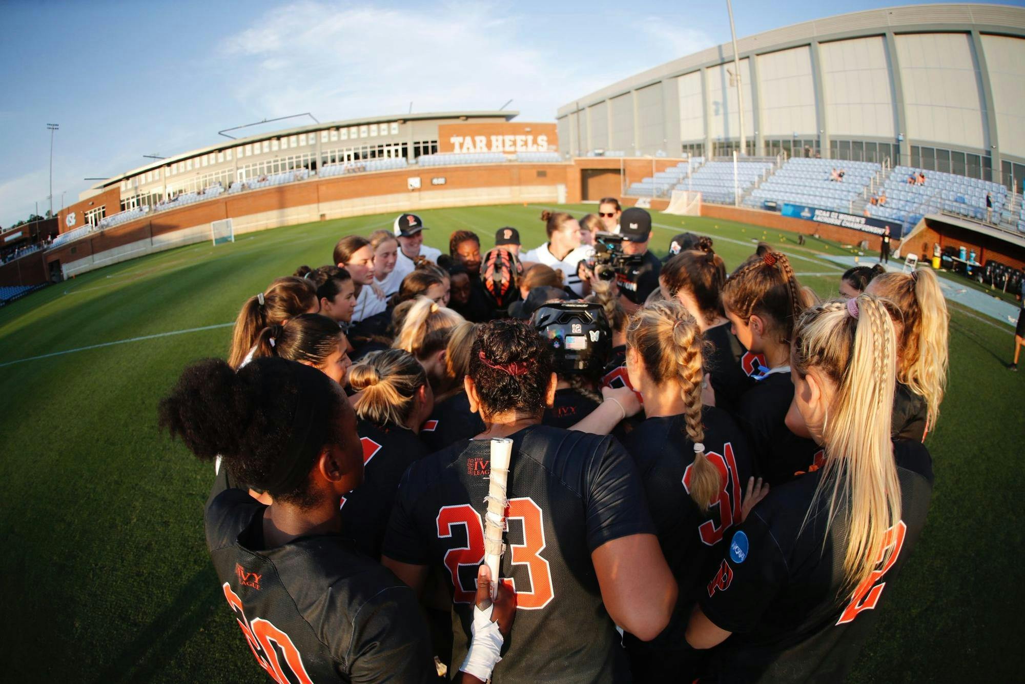 A group of women wearing black jerseys huddled up on a grass field following the conclusion of a lacrosse game.