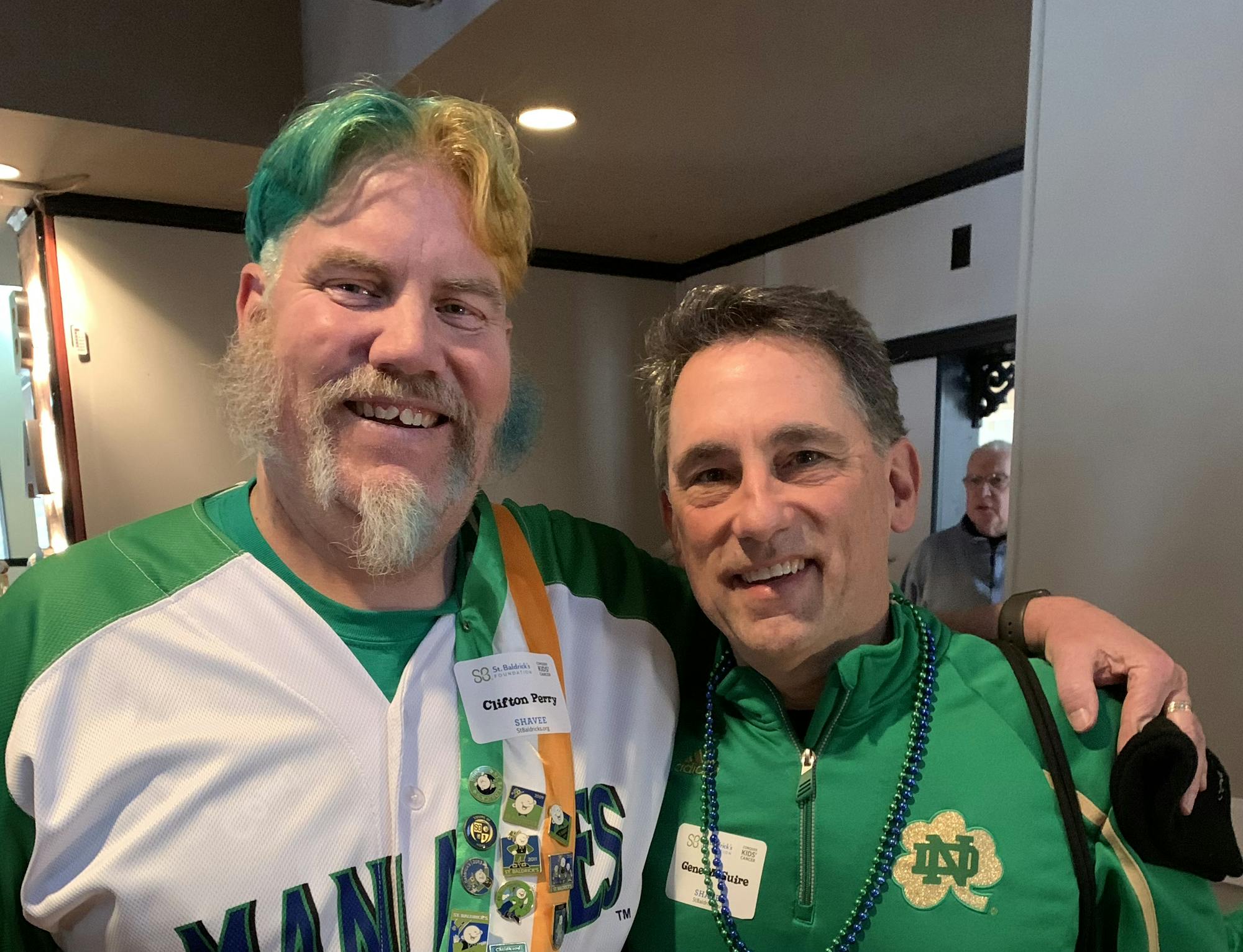 Two men pose for a photo. The man on the left has the left side of his hair dyed green and the right side dyed yellow. 