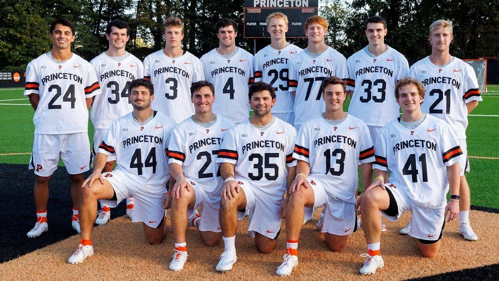 Thirteen men in orange and white Princeton uniforms pose for photo on the lacrosse field.