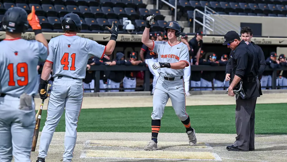 Baseball player in grey uniform runs to homeplate where two teammates are waiting.