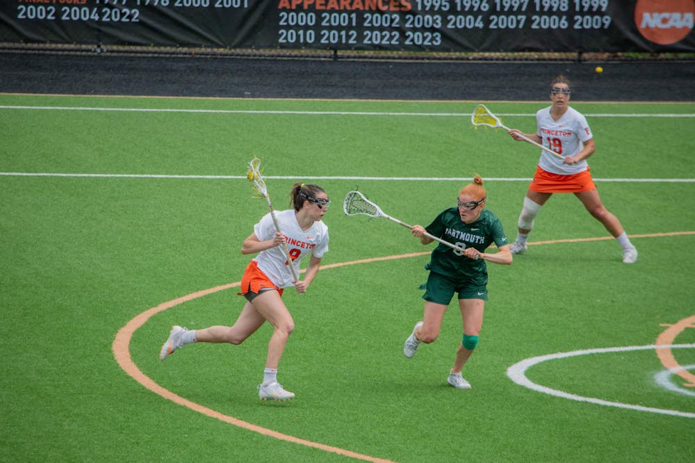 A woman running on a field with a lacrosse stick in her hand while a defender in green guards her.