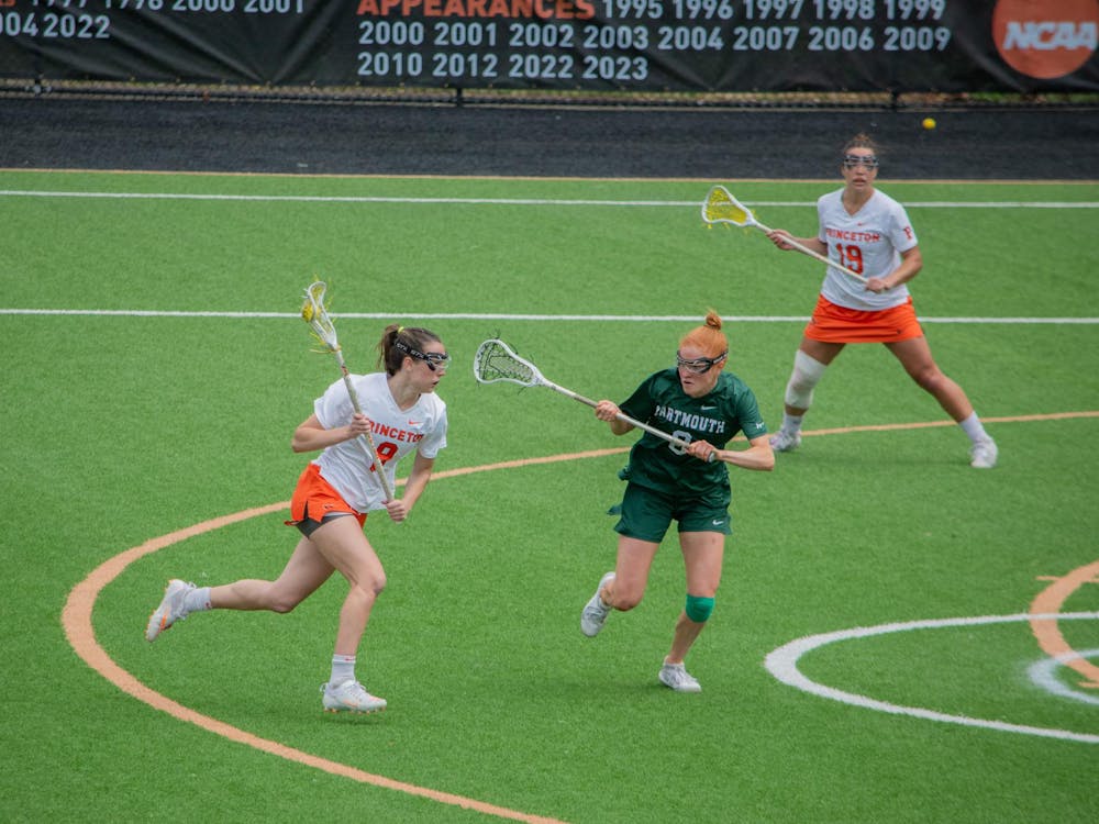 A woman running on a field with a lacrosse stick in her hand while a defender in green guards her.