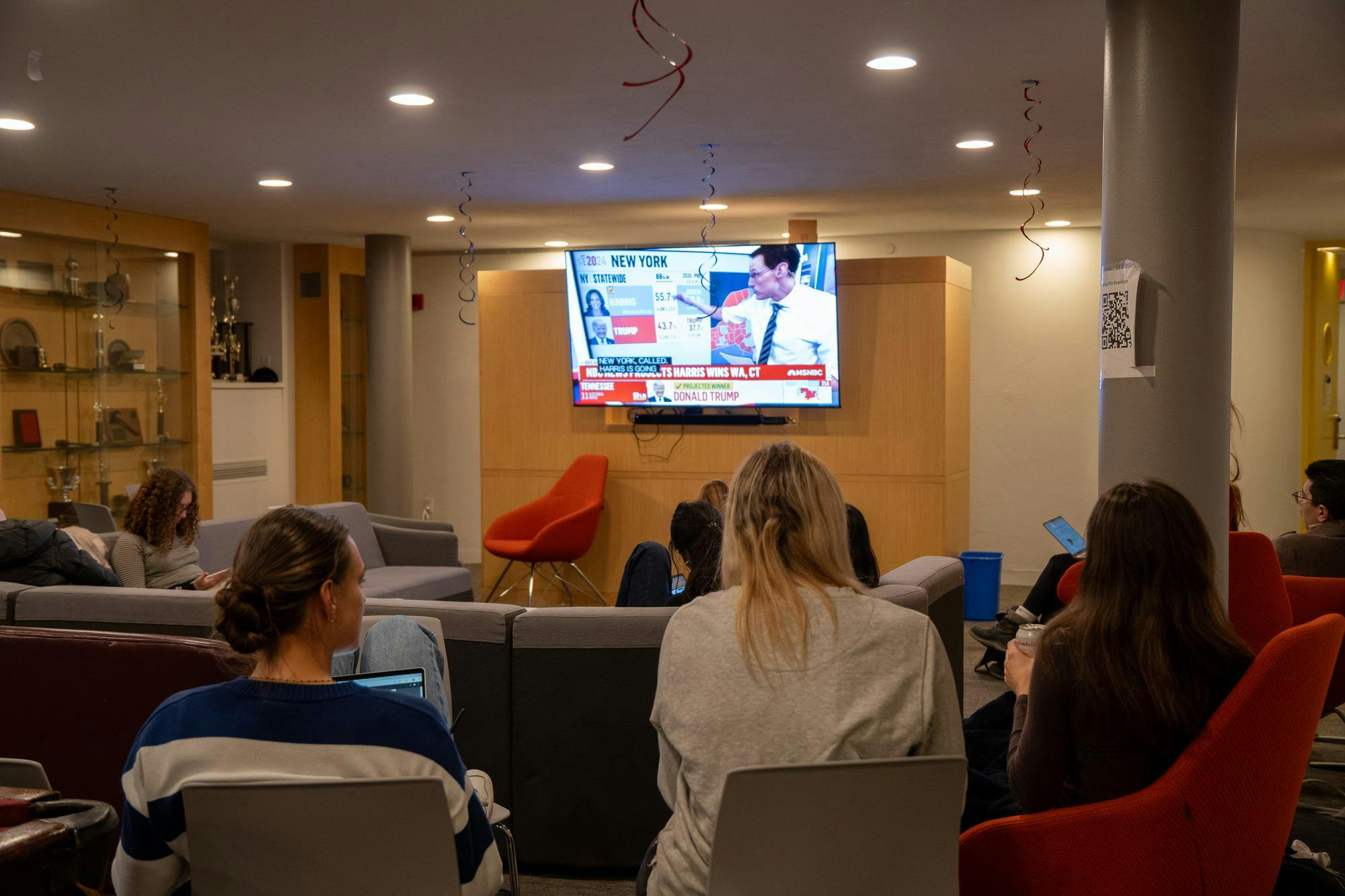 Three students watch MSNBC on a TV in the distance.