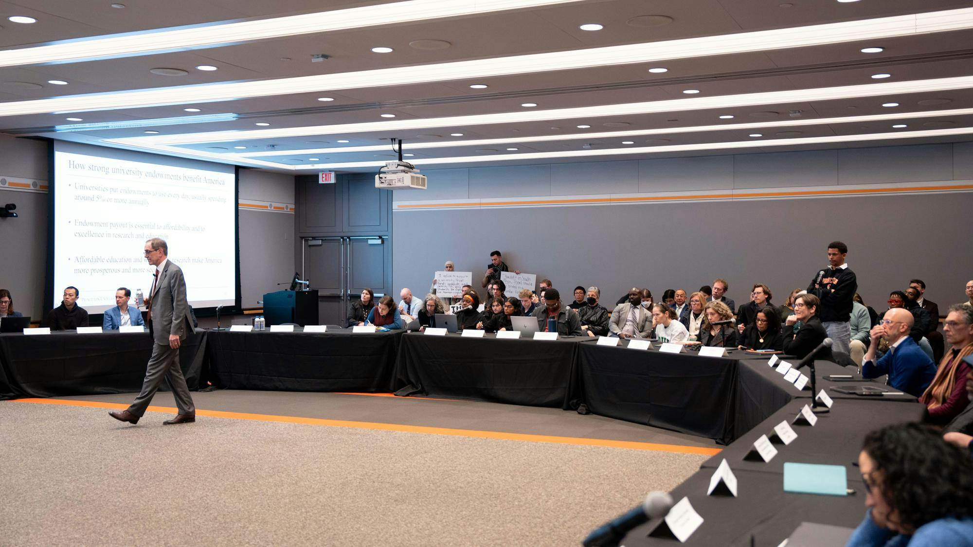 Man in gray suit and glasses walks in opposite direction of tables with black tableclothes and crowd of people.