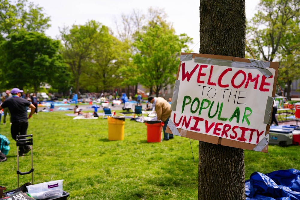 People sit on a green lawn. In the foreground, a sign tied to a tree reads "WELCOME TO THE POPULAR UNIVERSITY."