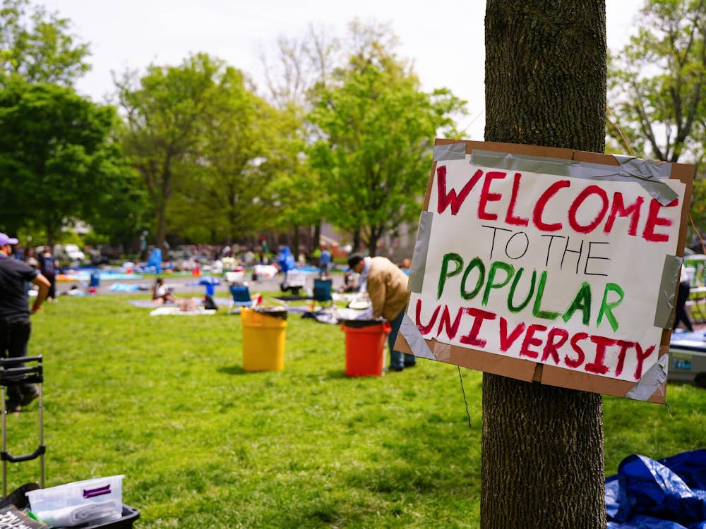 People sit on a green lawn. In the foreground, a sign tied to a tree reads "WELCOME TO THE POPULAR UNIVERSITY."