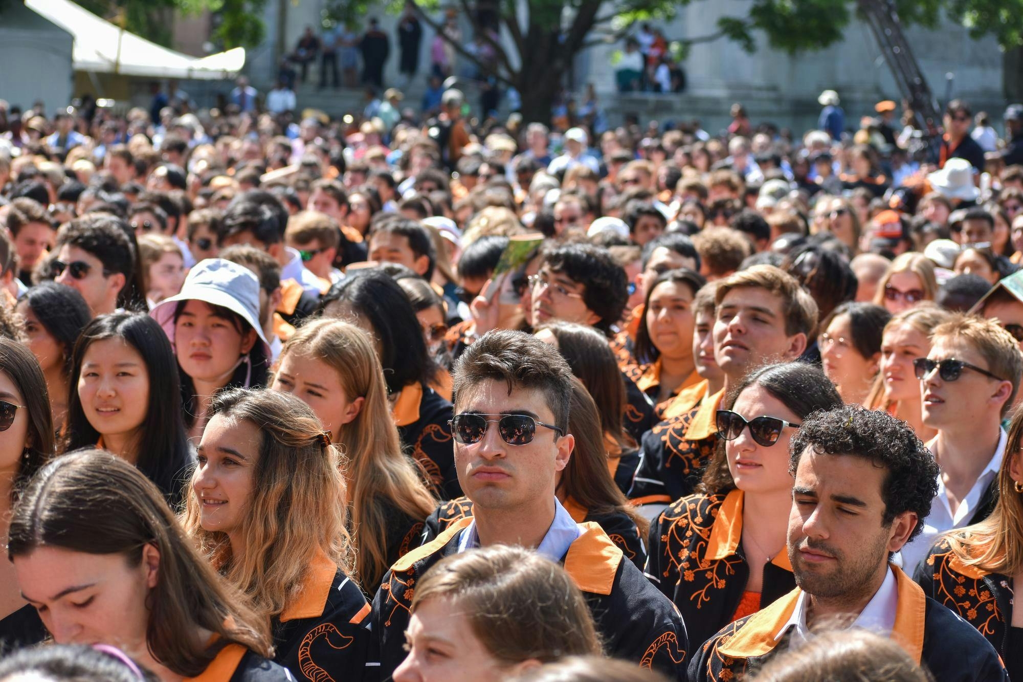 Crowd of people in orange and black jackets sit outside and listen to a speaker out of frame.