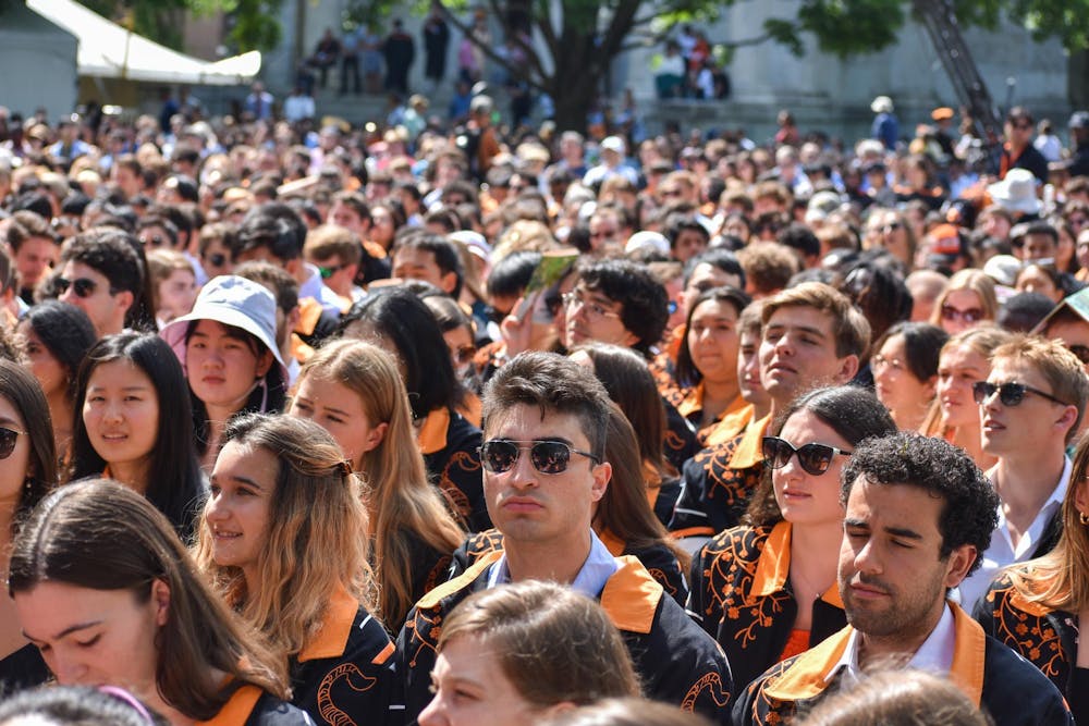 Crowd of people in orange and black jackets sit outside and listen to a speaker out of frame.