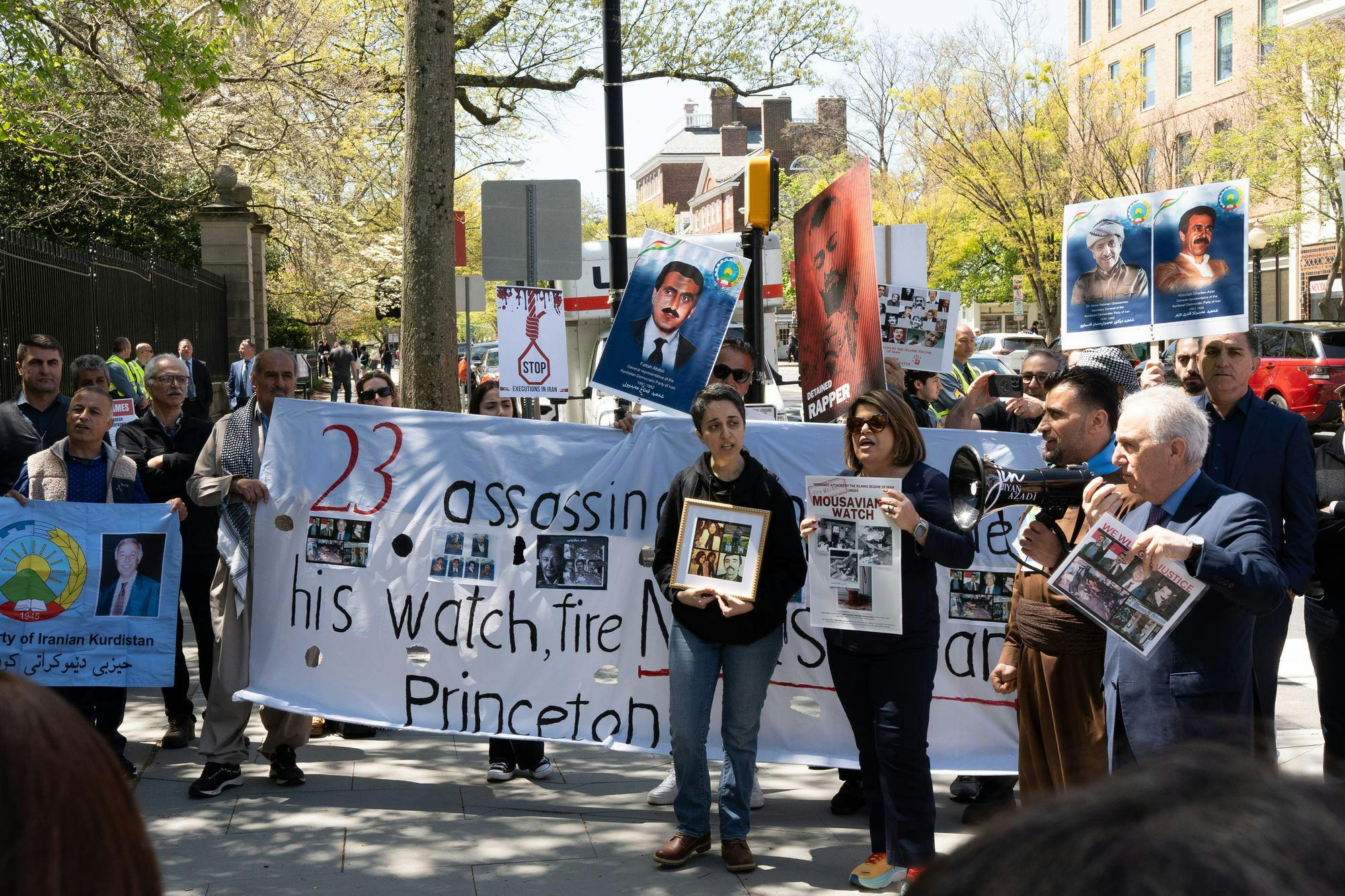 A group of protesters stand in a semi-circle in front of a large black gate. Some hold up posters with photographs of men. 
