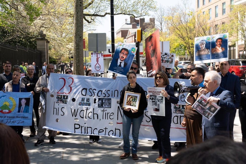 A group of protesters stand in a semi-circle in front of a large black gate. Some hold up posters with photographs of men. 