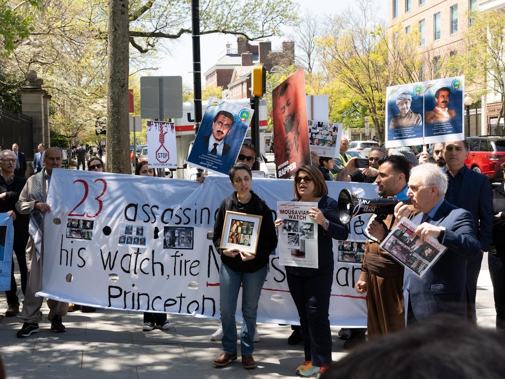 A group of protesters stand in a semi-circle in front of a large black gate. Some hold up posters with photographs of men.