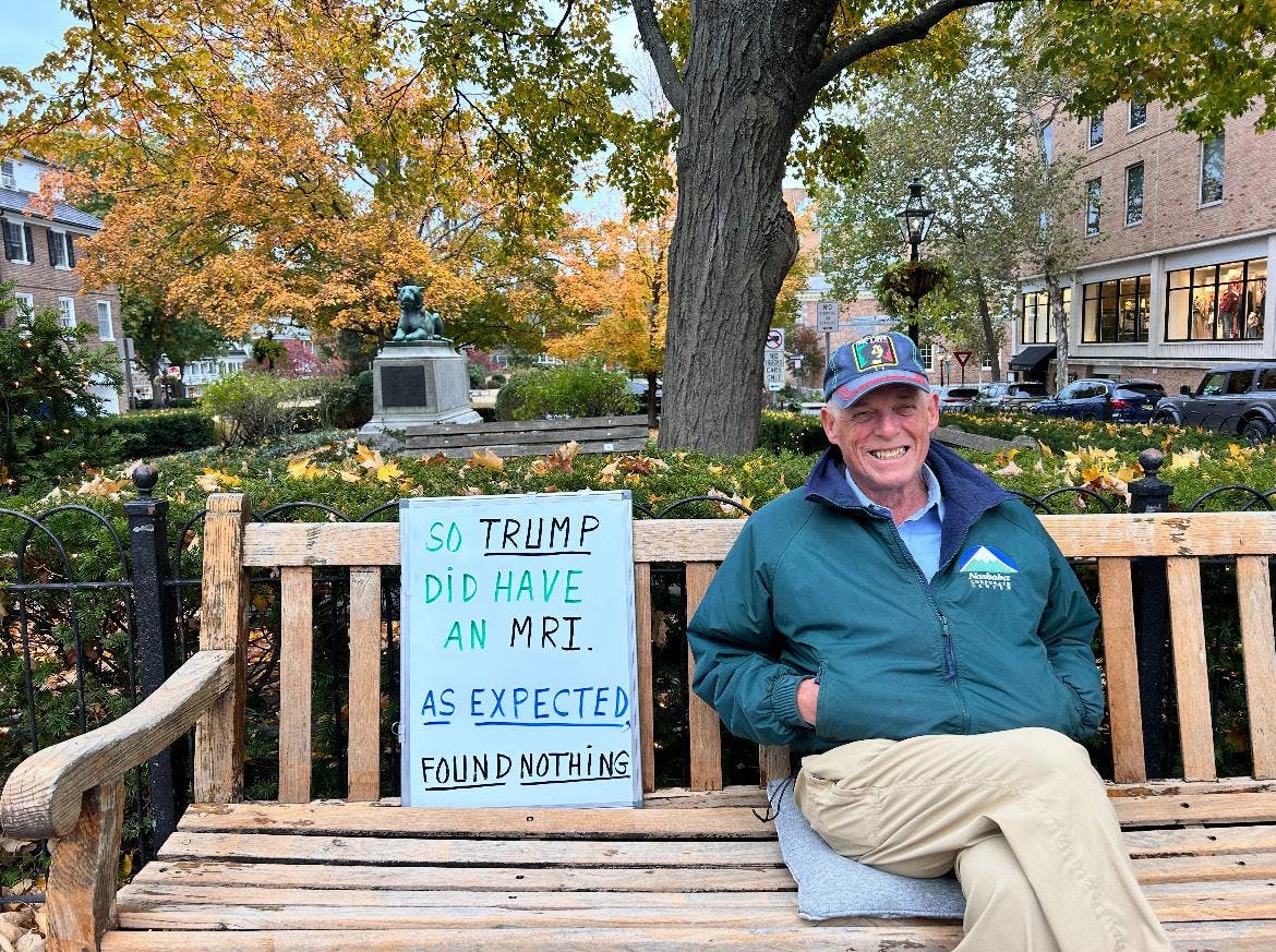 An old man with a green jacket sits on a bench with a sign, with a tree in the background.
