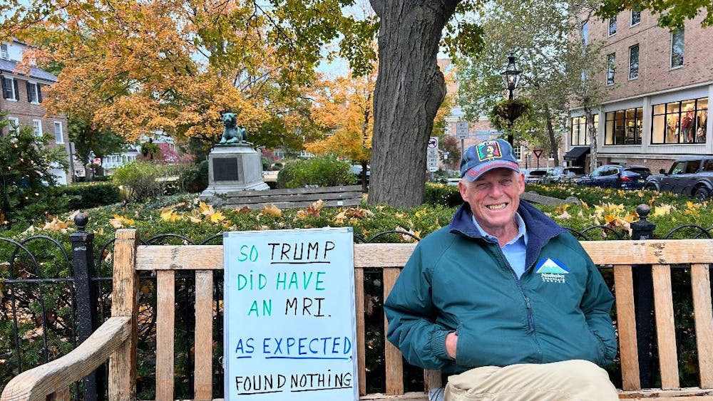 An old man with a green jacket sits on a bench with a sign, with a tree in the background.