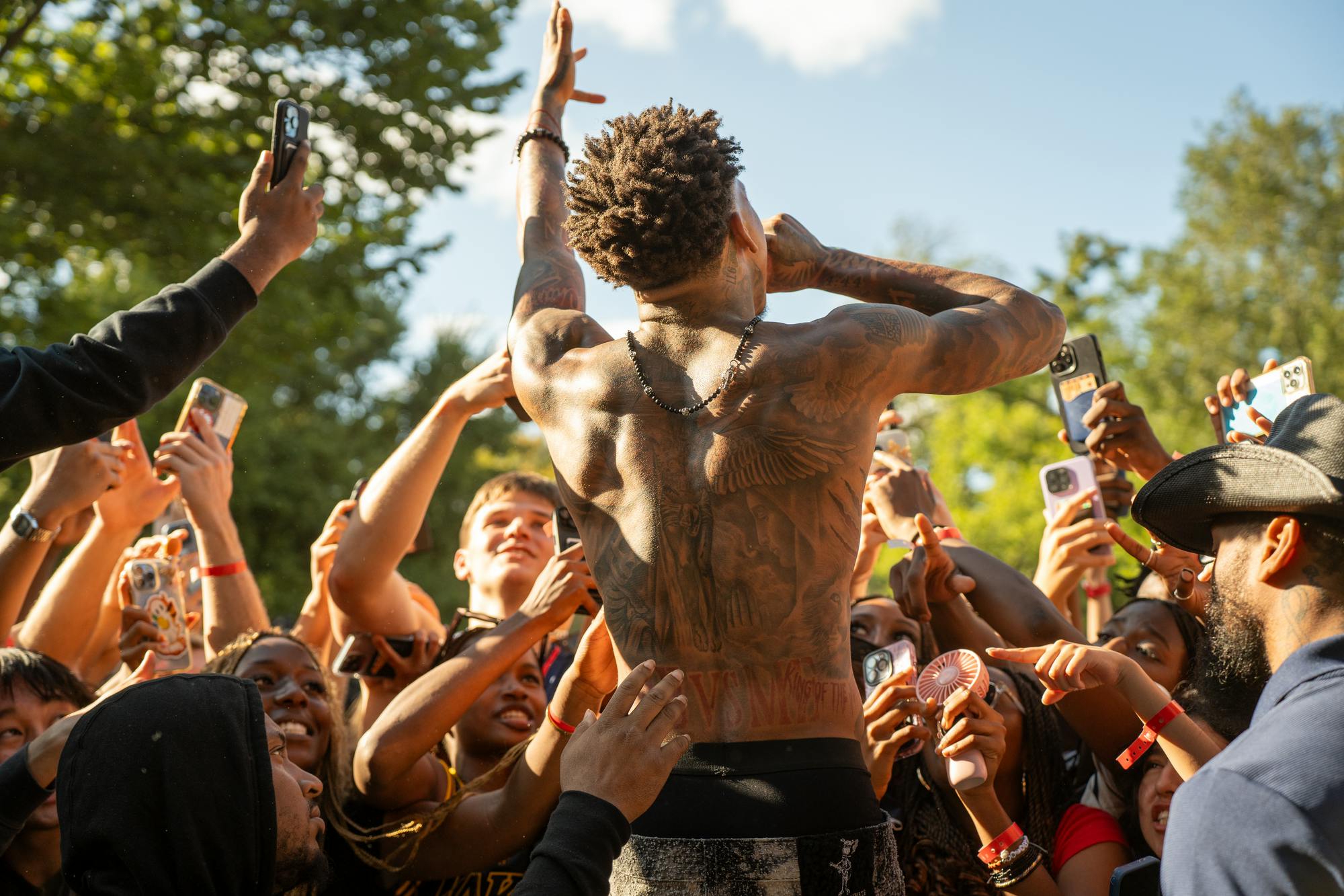 Man with exposed back and tattoos (NLE Choppa) speaks into mic in front of student crowd.